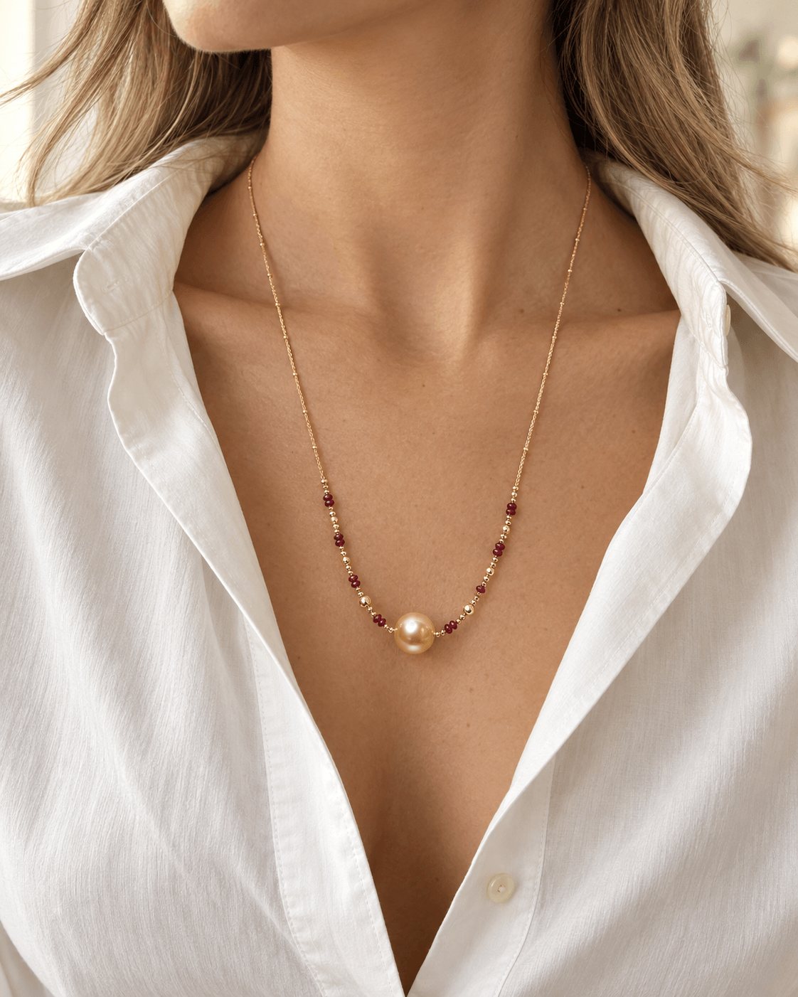A close-up of a woman wearing a delicate gold necklace with a single pearl pendant and small dark red bead accents, styled with a white button-down shirt.