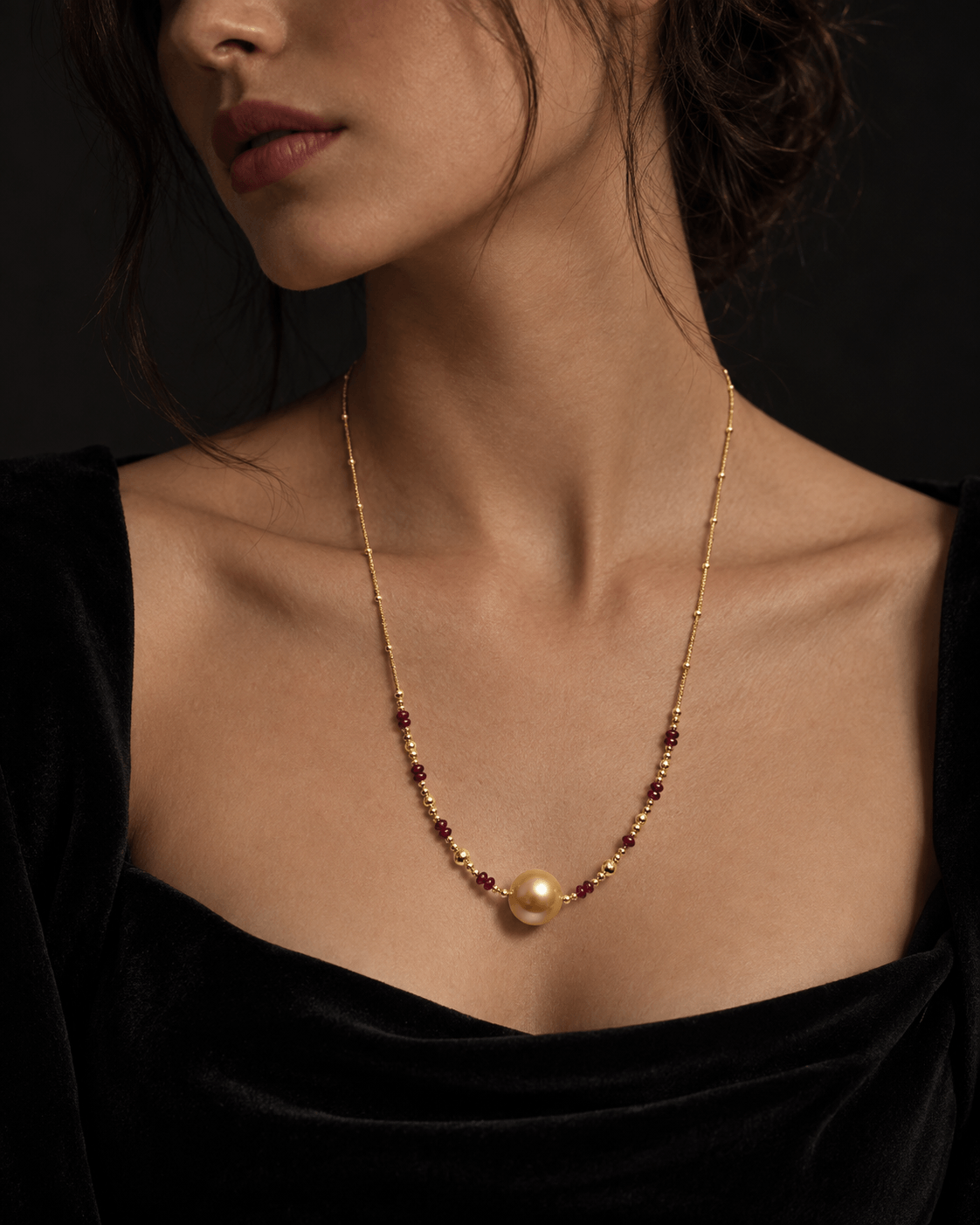 A close-up of a model wearing a delicate gold chain necklace with a single large pearl pendant and small dark red bead accents, styled with a black velvet top against a dark background.