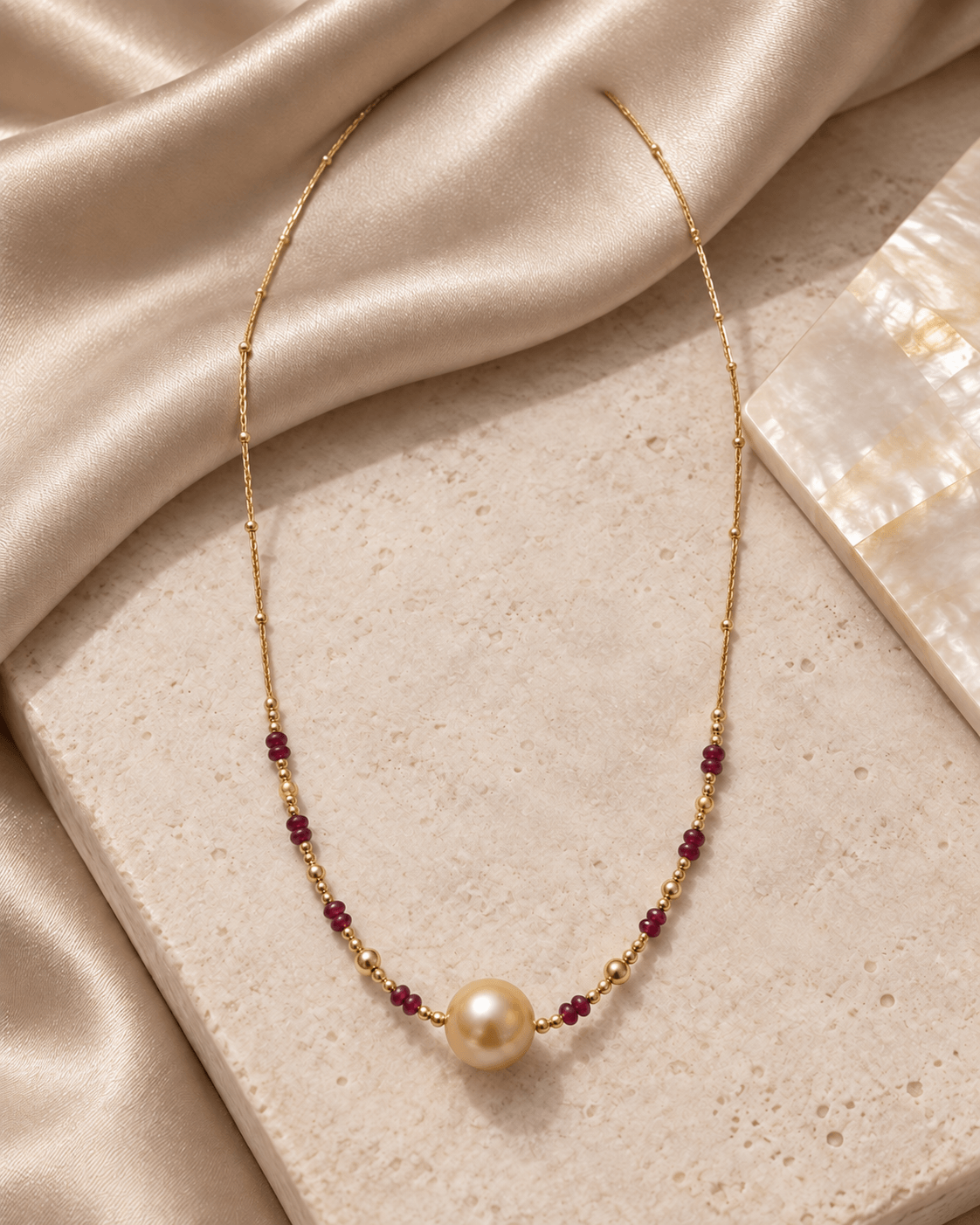 A delicate gold necklace featuring a single cream pearl at the center, flanked by small dark red gemstone beads and gold spacer beads, displayed on a textured stone slab with draped champagne silk.