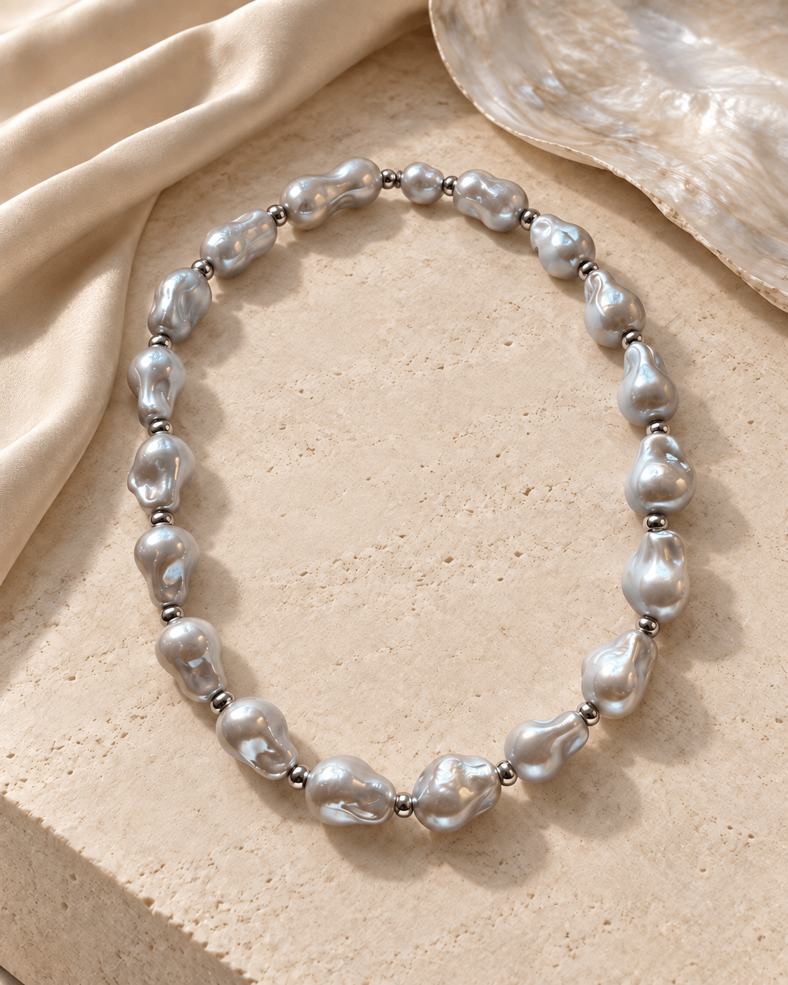 A baroque pearl necklace with irregular silver-gray pearls separated by small metallic spacer beads, arranged in an oval on a textured beige stone surface with draped satin and a shell in the background.