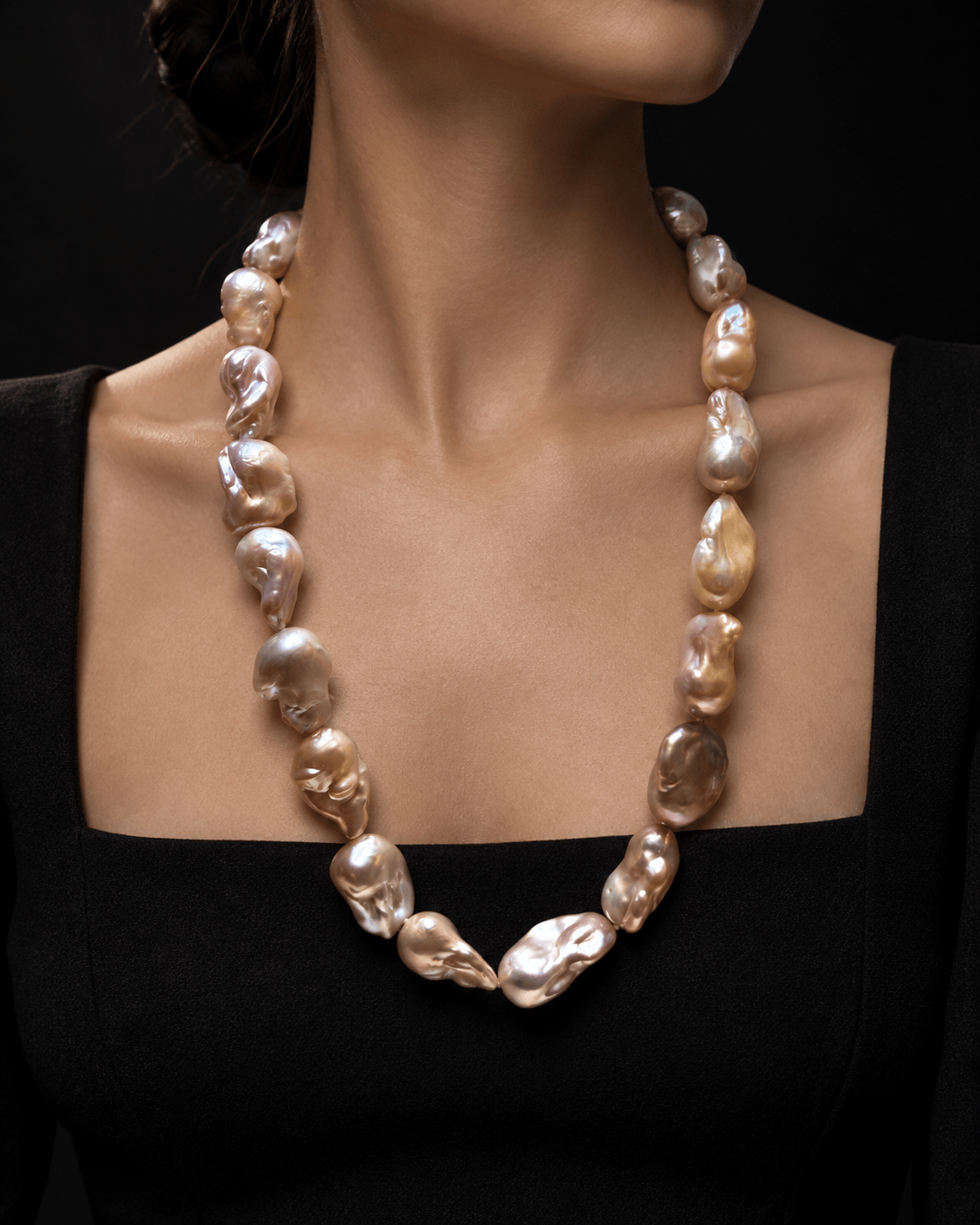 A close-up of a model wearing a baroque pearl necklace. The necklace features irregular, polished pearls in shades of champagne, beige, and soft gold, arranged in a classic circle around the neck.
