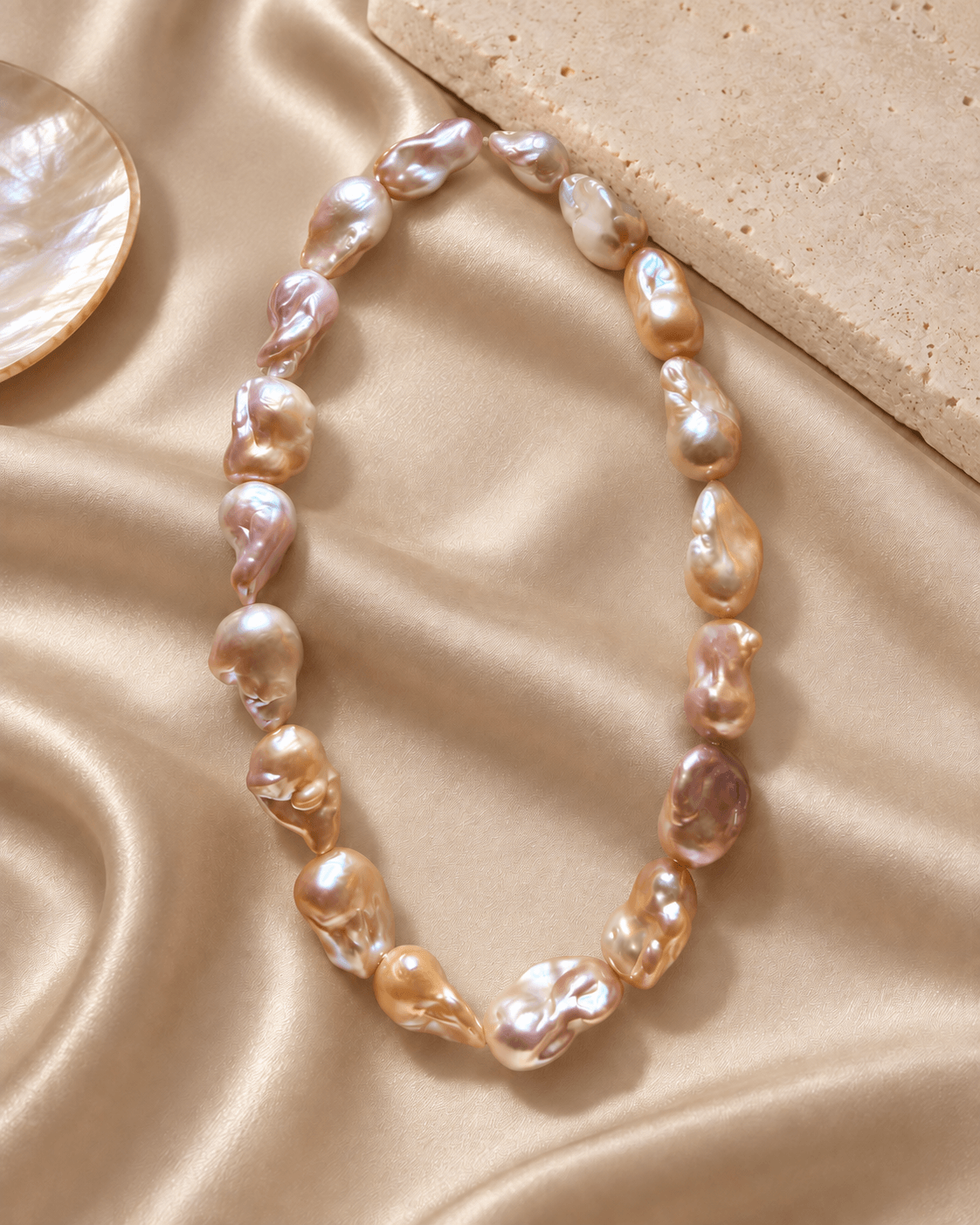 A baroque pearl necklace with irregular, lustrous pearls in champagne, gold, and soft pinkish-beige tones, arranged on draped champagne satin fabric with a stone prop.