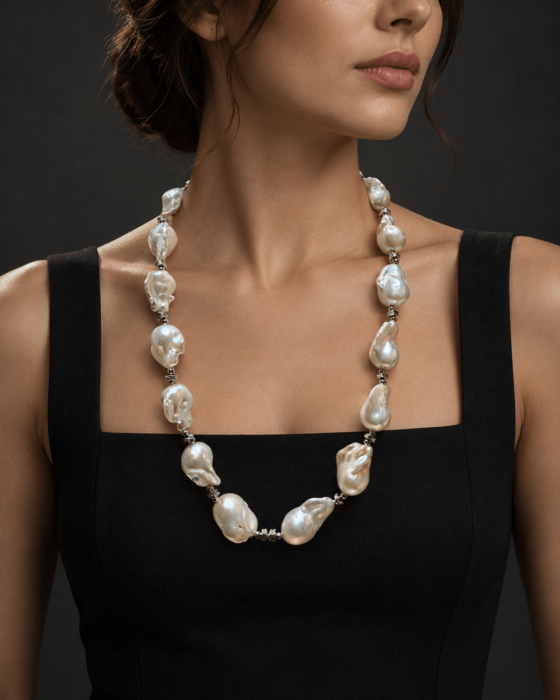 A close-up of a model wearing a baroque pearl necklace with silver-toned metal spacers, paired with a black sleeveless top against a dark gray background.