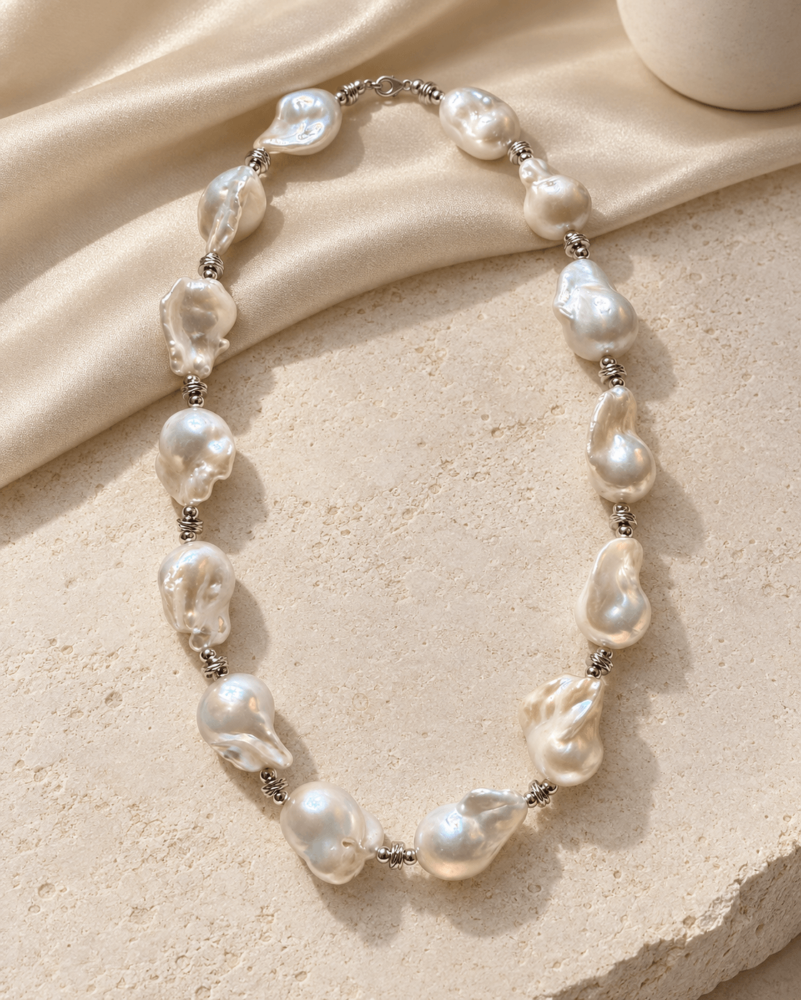 A baroque pearl necklace with irregular, lustrous off-white pearls separated by small silver spacer beads, arranged on a textured beige stone surface with draped satin fabric.