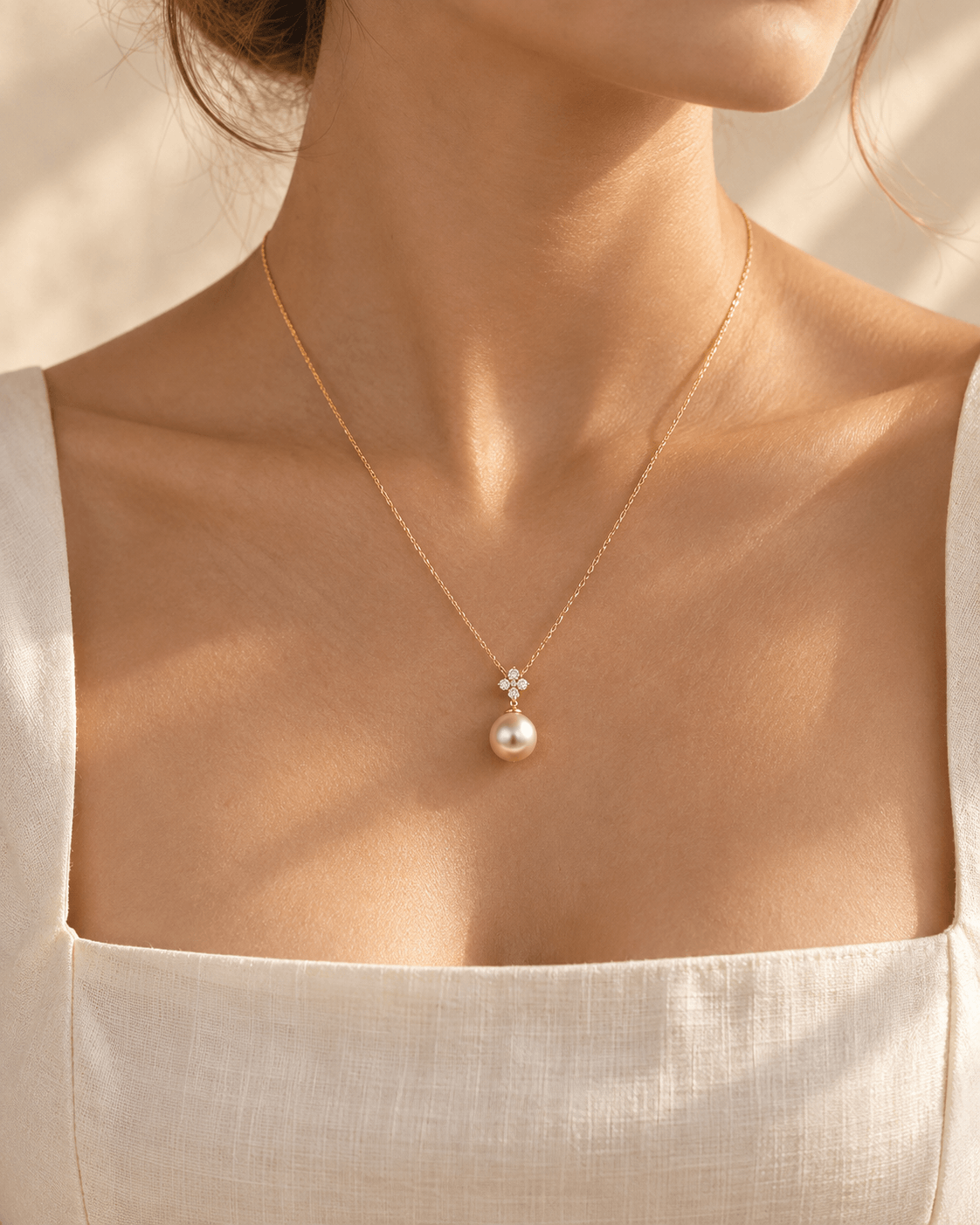 A close-up of a woman wearing a delicate gold pendant necklace with a teardrop pearl and a small diamond cluster, styled with a cream linen top.
