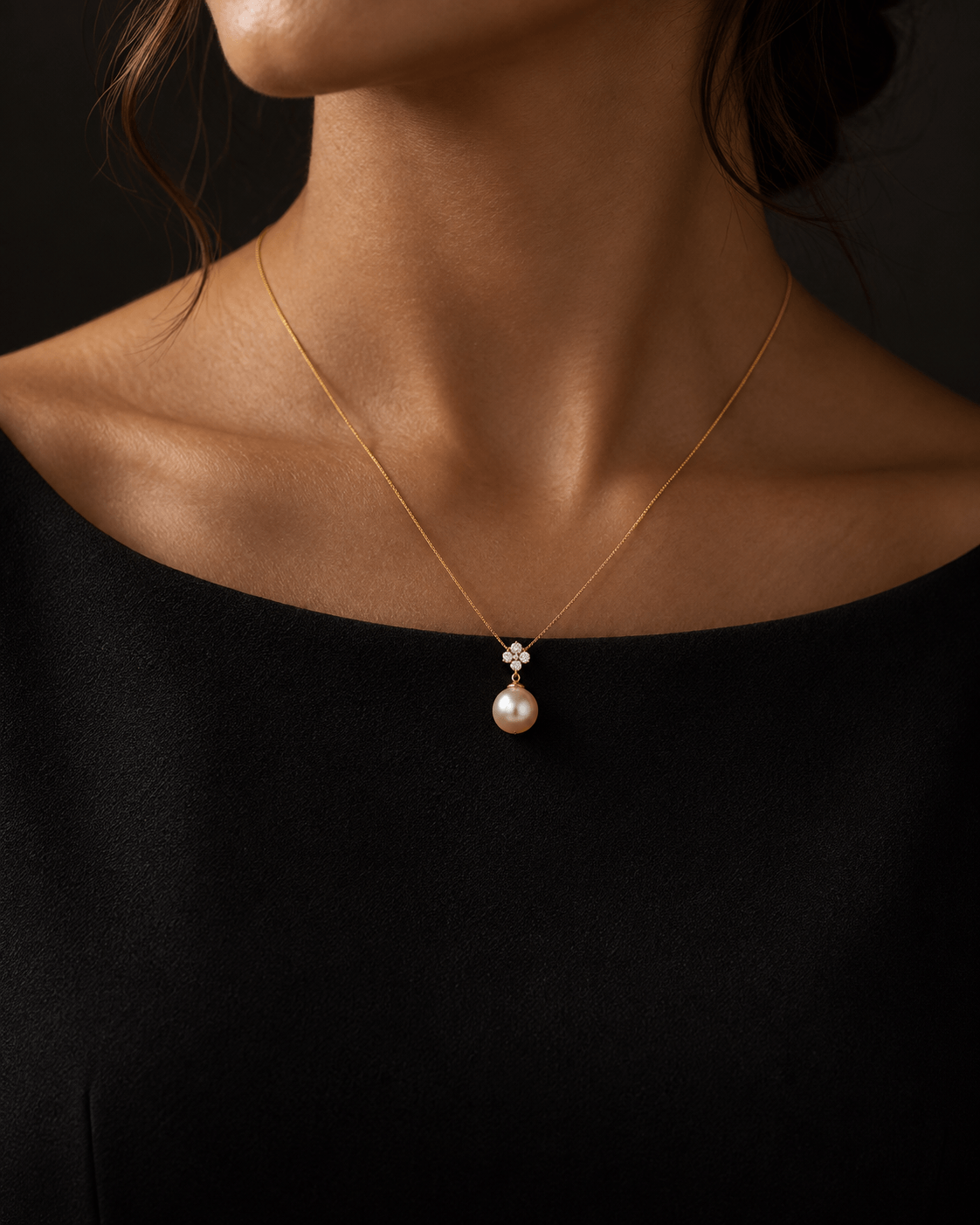 A close-up of a model wearing a delicate gold chain necklace with a pear-shaped pearl pendant set in a small diamond cluster, styled with a black top against a dark background.