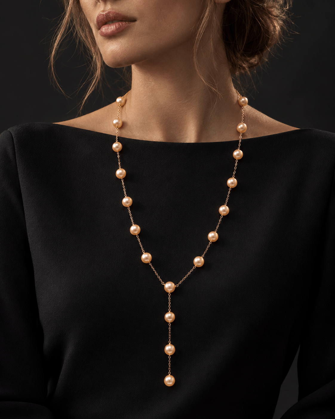 A woman wearing a rose gold Y-shaped pearl necklace over a black top, shown from the chest up against a dark background.