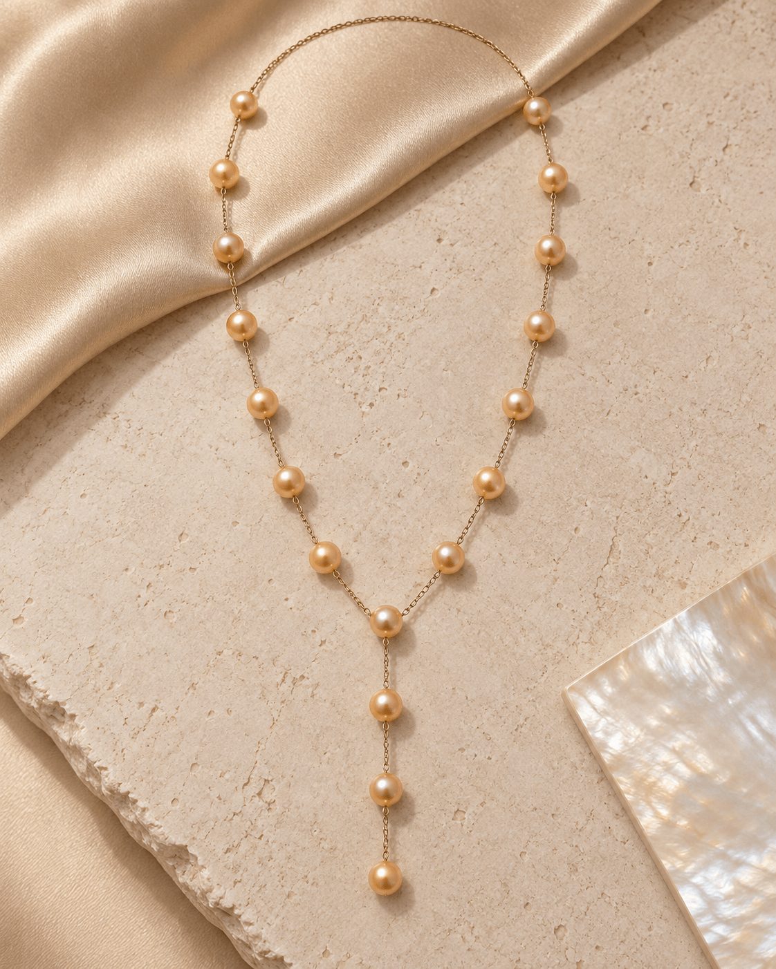A delicate gold Y-necklace featuring small round champagne-colored pearls, styled on a textured beige stone surface with draped champagne silk and a piece of mother-of-pearl paper.