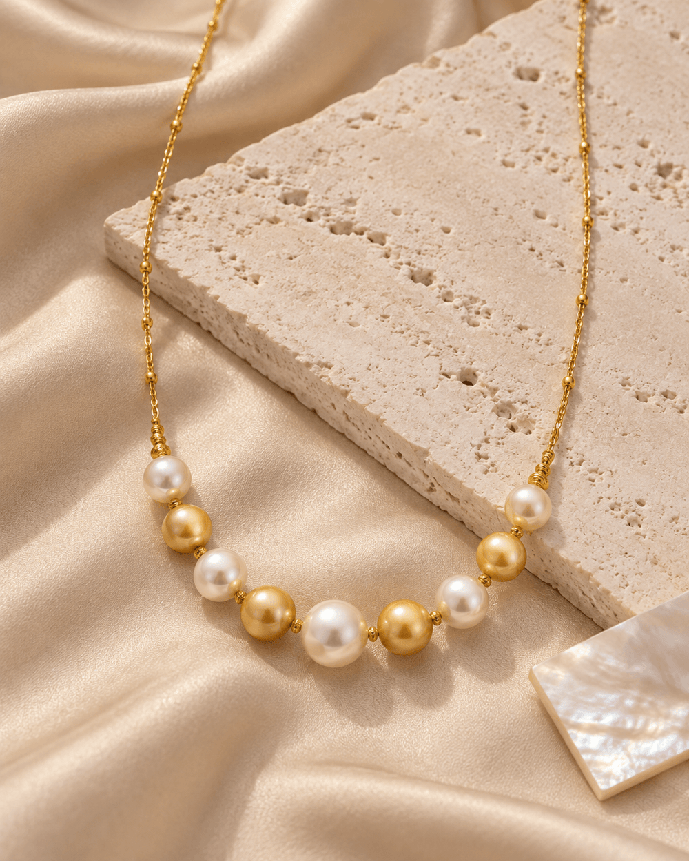 A gold chain necklace featuring a row of alternating white and gold freshwater pearls, styled on draped champagne satin fabric with a textured stone block.