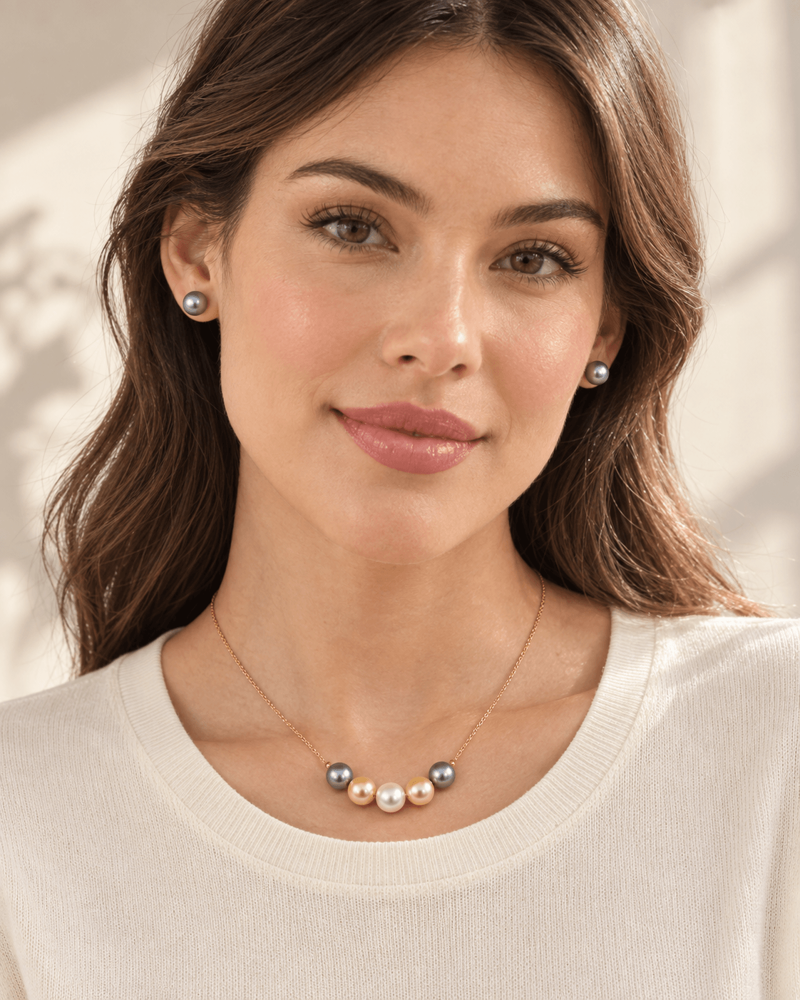 A woman wearing a delicate gold necklace with a row of small pearls and gray-blue beads, along with matching pearl stud earrings, shown in a warm, softly lit setting.
