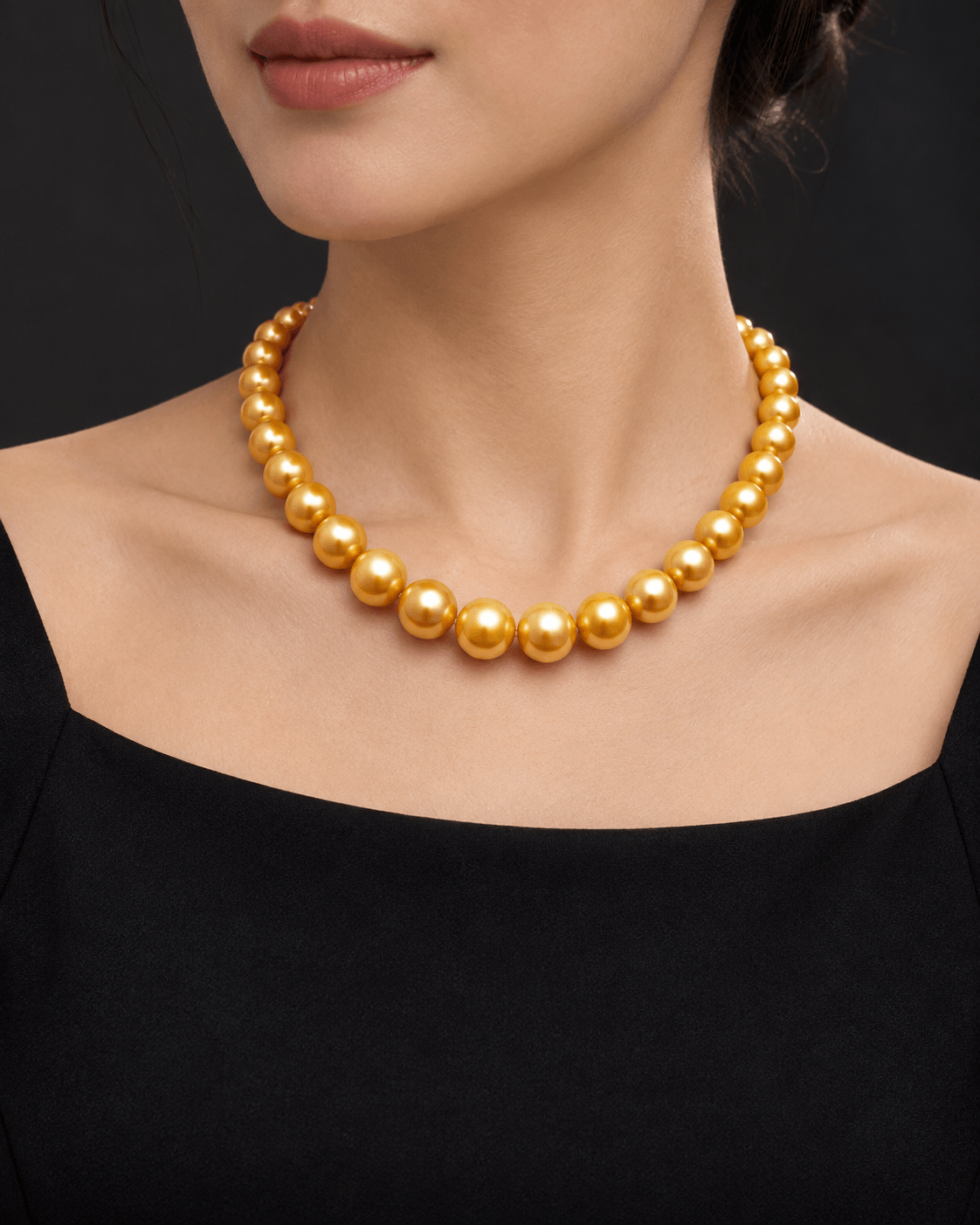 A close-up of a model wearing a gold beaded pearl necklace with large, polished round beads, paired with a black top against a dark background.
