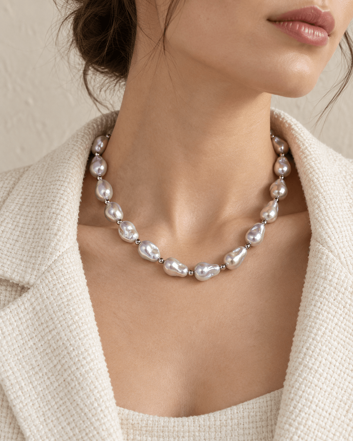 A close-up of a woman wearing a silver-toned necklace with irregular baroque pearls, styled with a textured cream blazer.