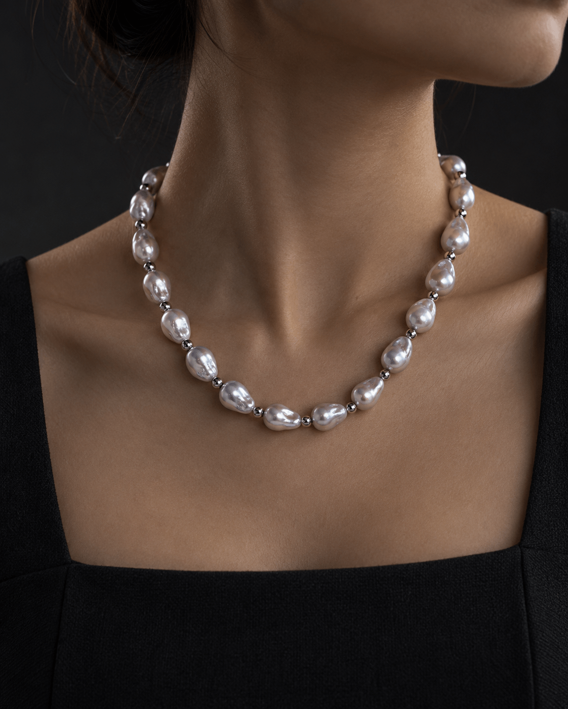 A close-up of a model wearing a silver-toned necklace with irregular baroque pearls, paired with a black top against a dark background.