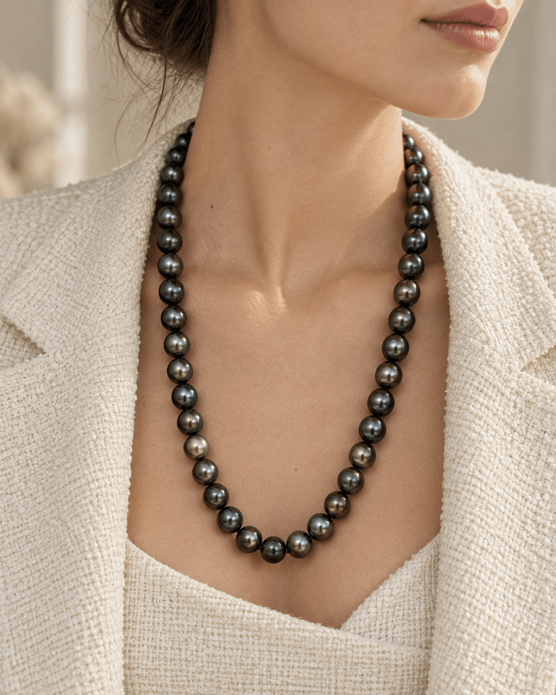 A close-up of a woman wearing a dark gray to black freshwater pearl necklace with a matte finish, styled with a textured cream blazer.