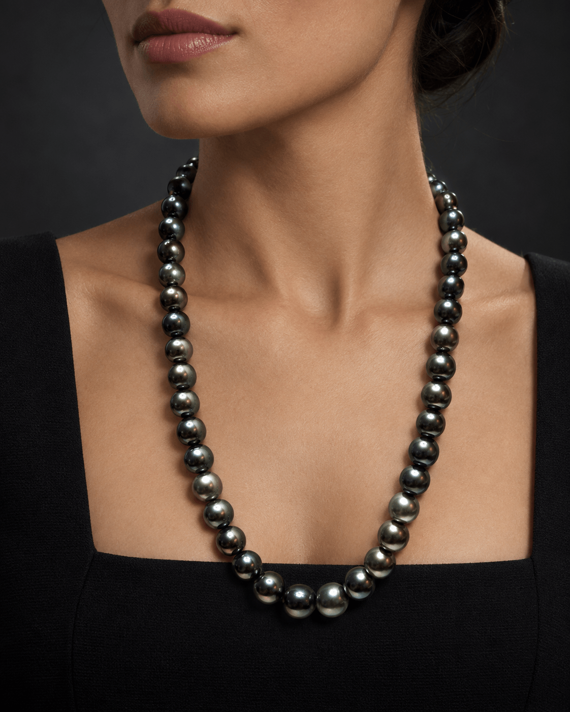 A close-up of a model wearing a dark gray to black freshwater pearl necklace with a square neckline and a black top, set against a dark gray background.