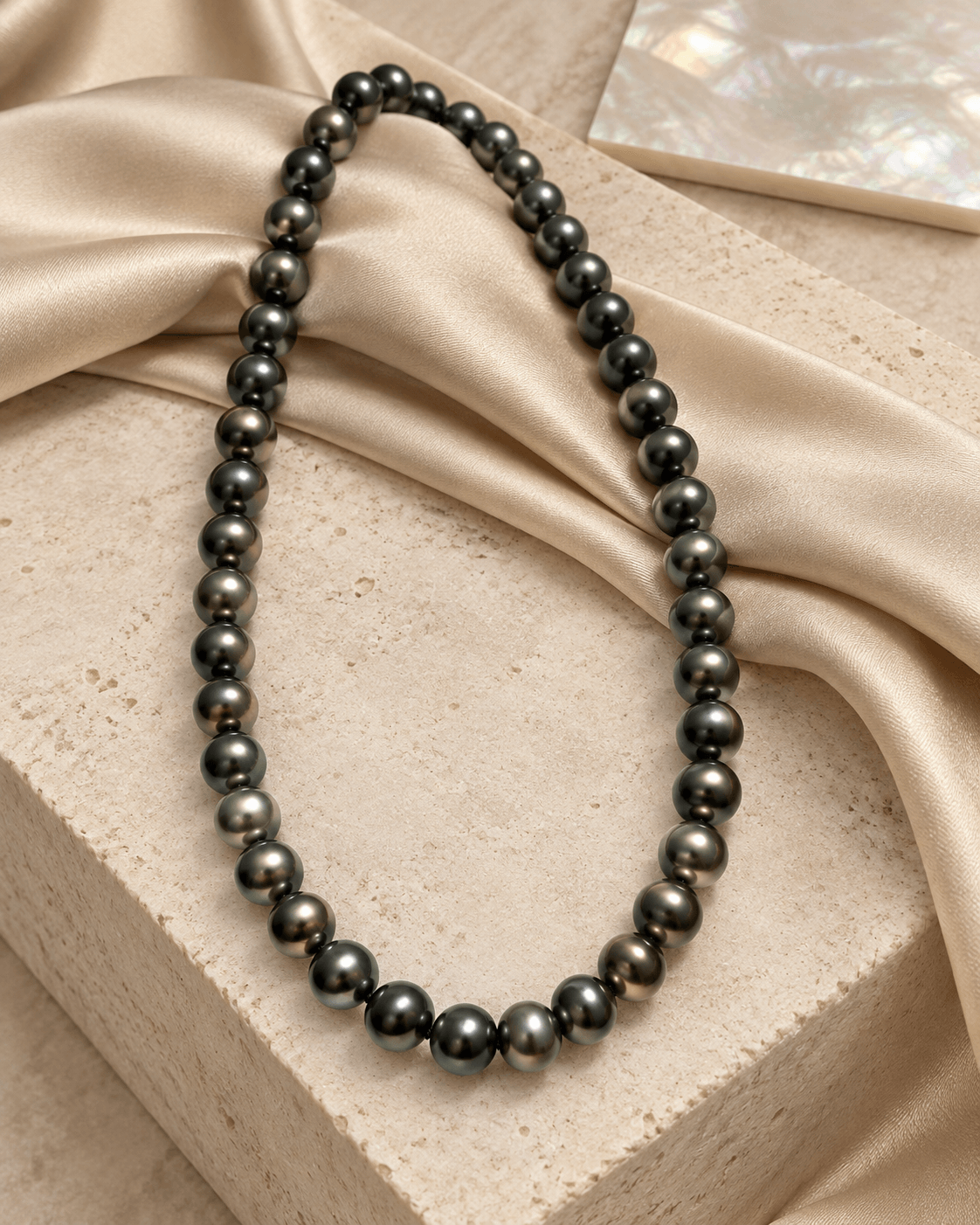 A dark gray pearl necklace draped elegantly over a beige stone block with beige satin fabric.