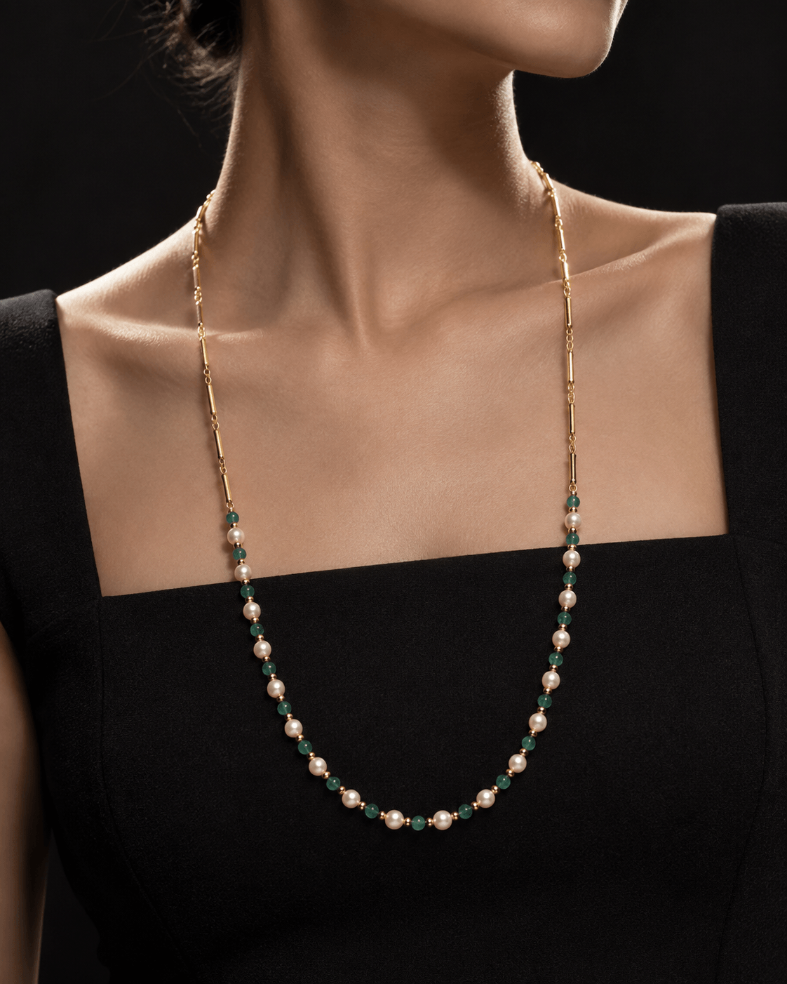 A close-up of a model wearing a delicate necklace with small white pearls, green emeralds, and gold-toned metal links, styled with a black square-neck top against a dark background.