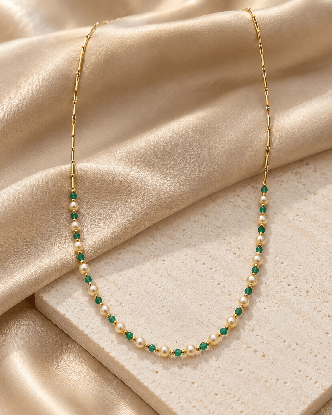 A delicate gold necklace featuring small white pearls and emerald green gemstone accents, draped elegantly over a beige textured stone slab and cream satin fabric.