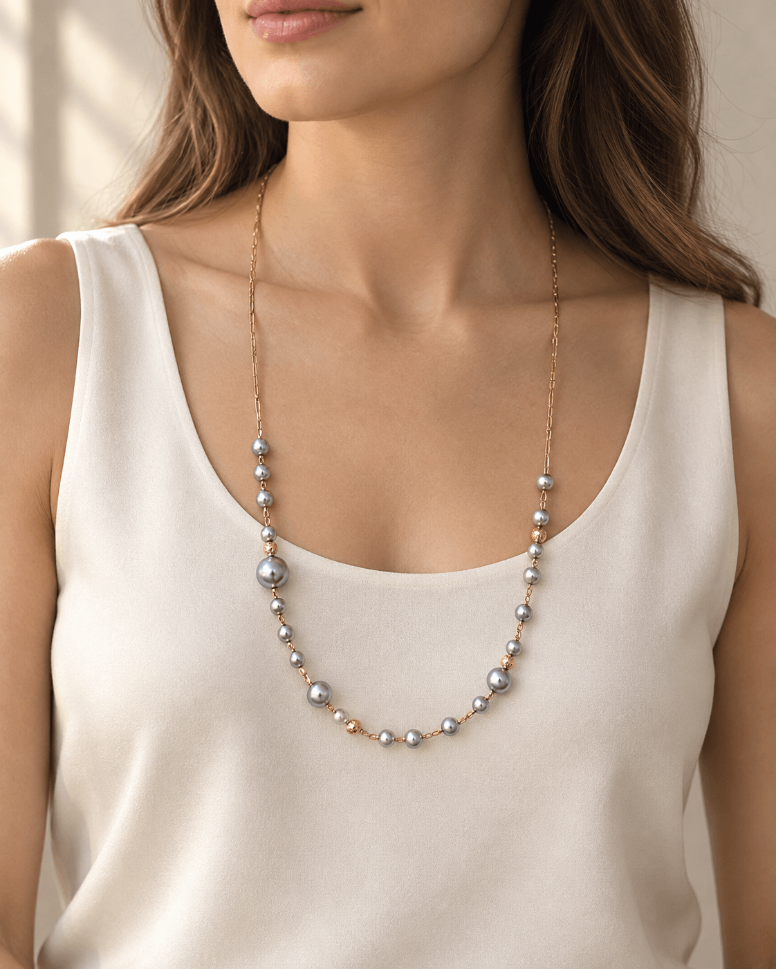 A woman wearing a delicate necklace with small gray pearls and rose gold accents, styled with a cream-colored tank top.