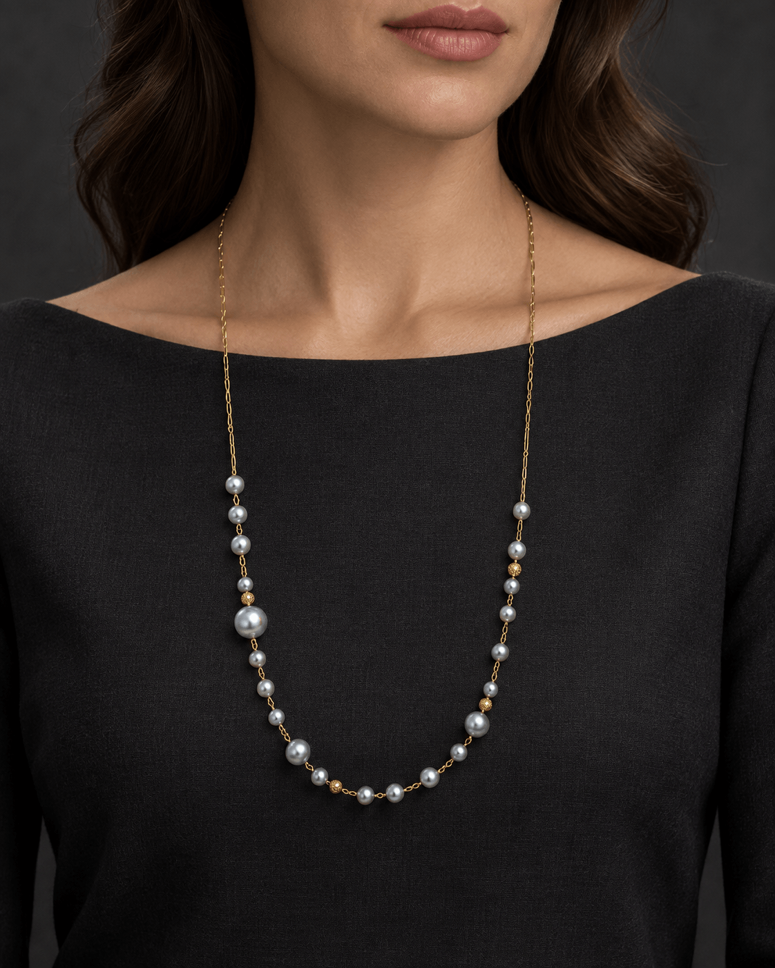 A close-up of a model wearing a delicate gold chain necklace with small gray pearls and a few gold spacer beads, styled with a black top against a dark gray background.