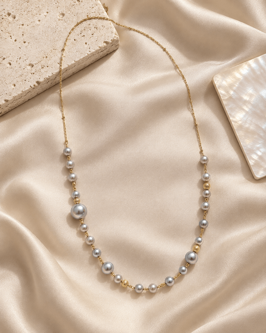 A delicate gold chain necklace featuring a graduated strand of gray pearls, styled on a draped beige silk fabric with decorative stone props.