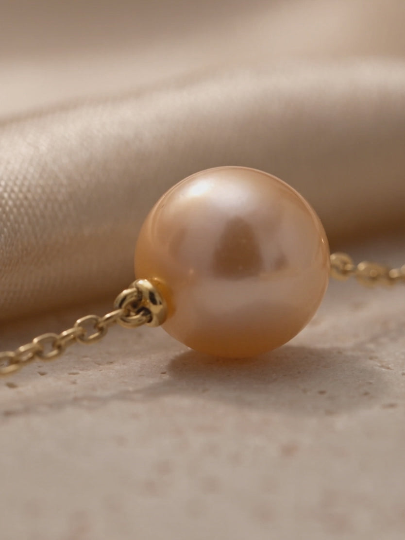 Anna Borjigin golden-peach pearl necklace detail showcasing a lustrous round pearl connected by a delicate gold chain on natural stone.