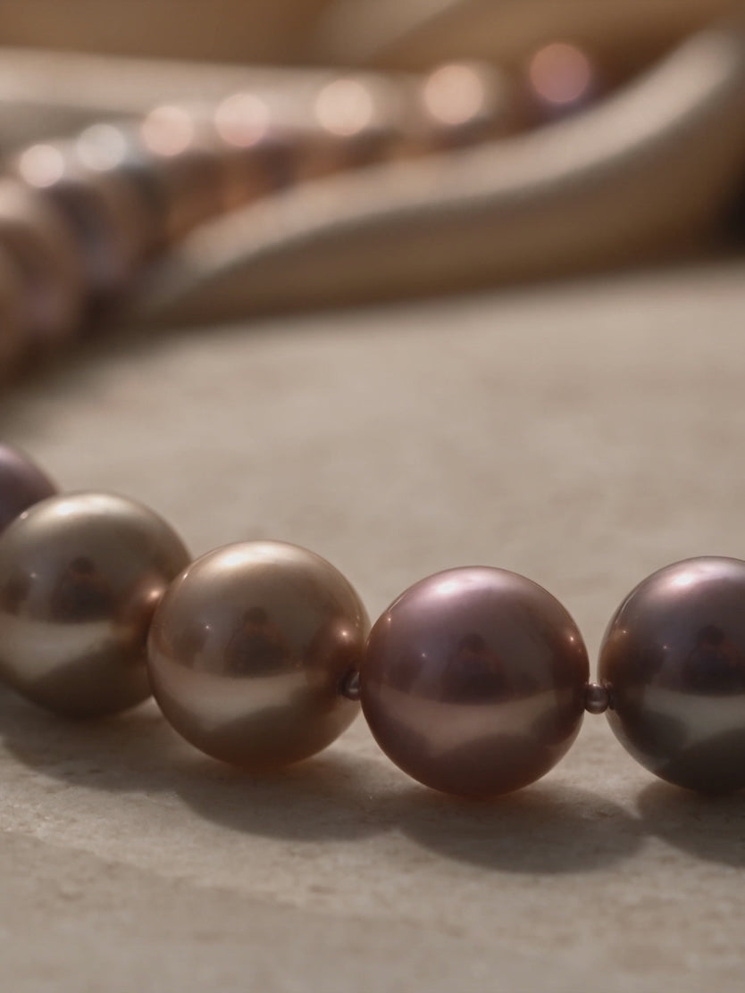 Close-up video of a lustrous multi-toned pearl necklace featuring bronze, gold, and mauve pearls on a warm neutral surface.