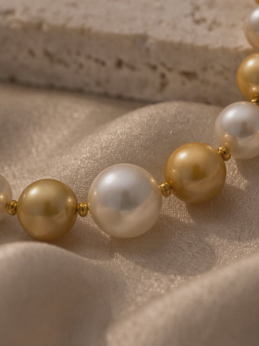Alternating white and golden South Sea style pearl necklace with gold bead spacers displayed on neutral fabric in warm lighting.