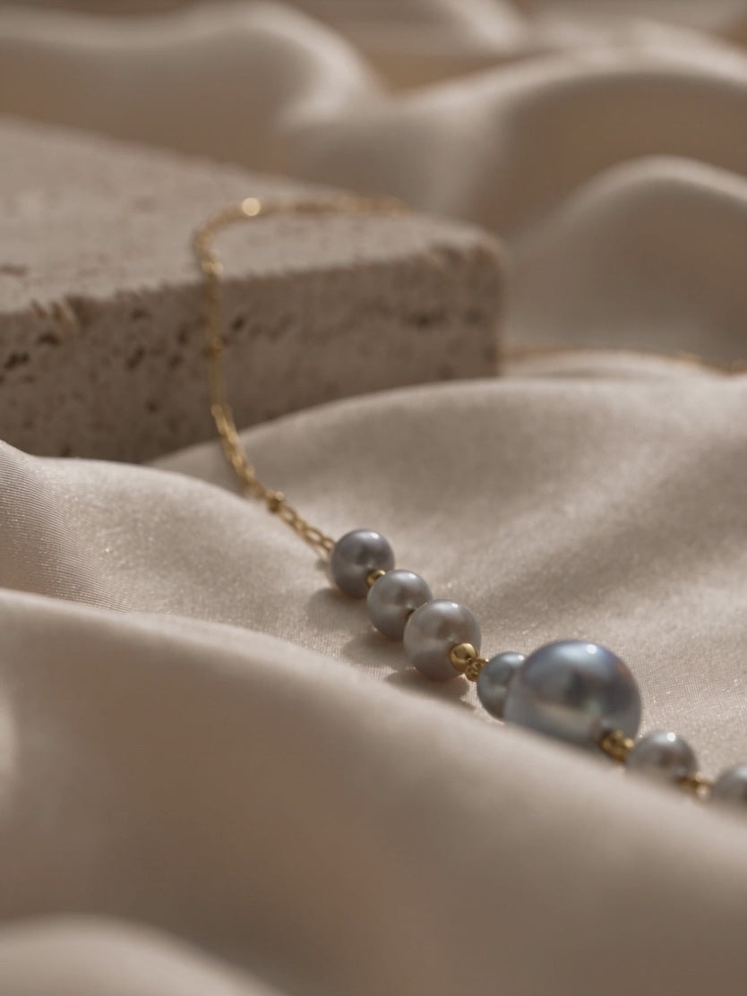 Delicate gold link necklace with graduated silver-grey pearls and gold bead accents displayed on champagne satin.